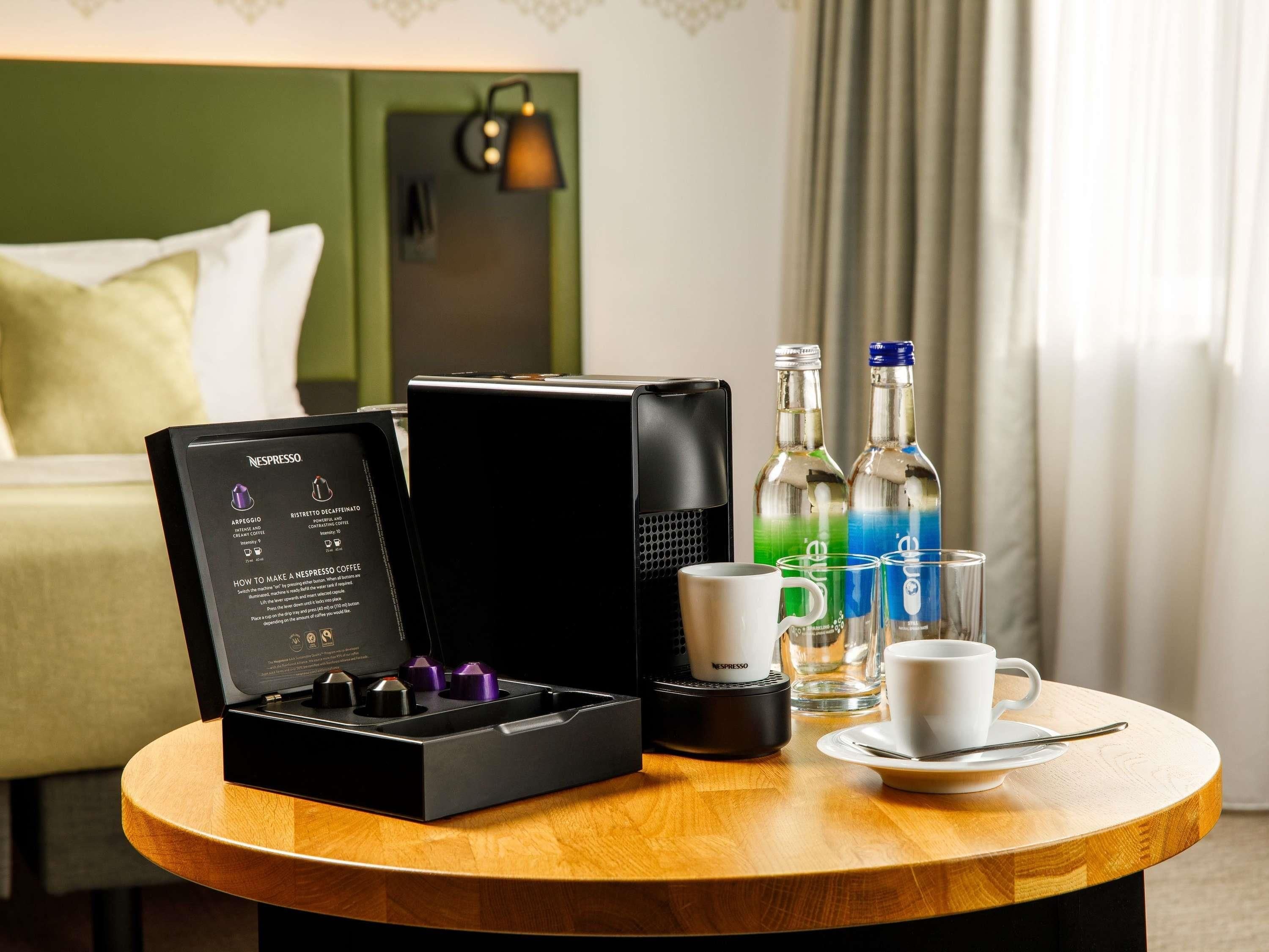 A Nespresso coffee maker, two bottles of water, a cup, and a saucer on a round wooden table in a hotel room.