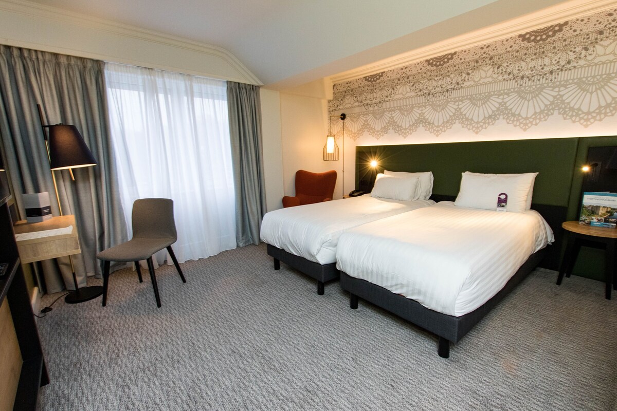 A hotel room with a double bed, a desk with a chair, a lamp, and a decorative wall piece.