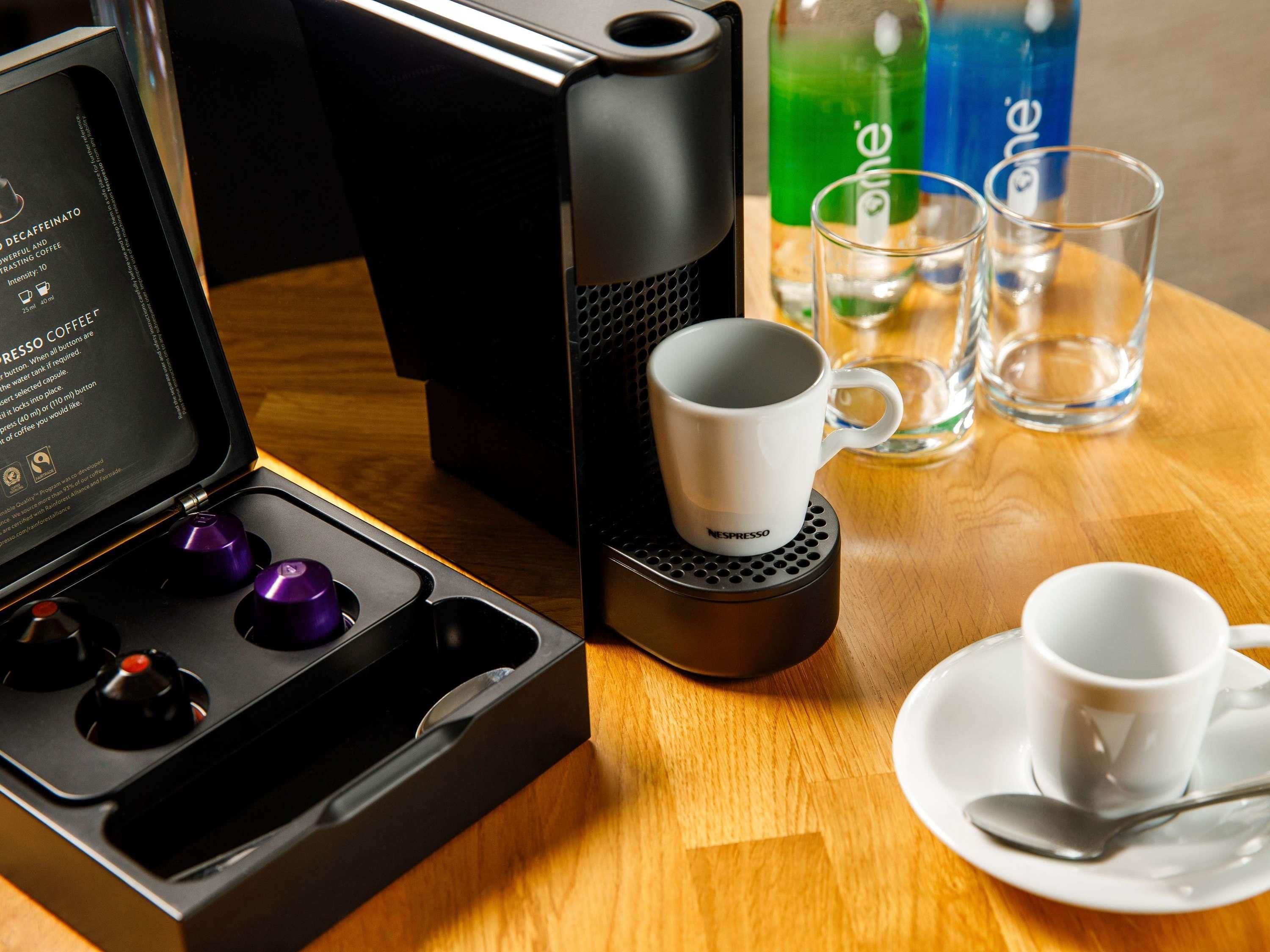 Een Nespresso koffieapparaat met een kop en schotel, een glas water en een waterfles op een houten tafel.