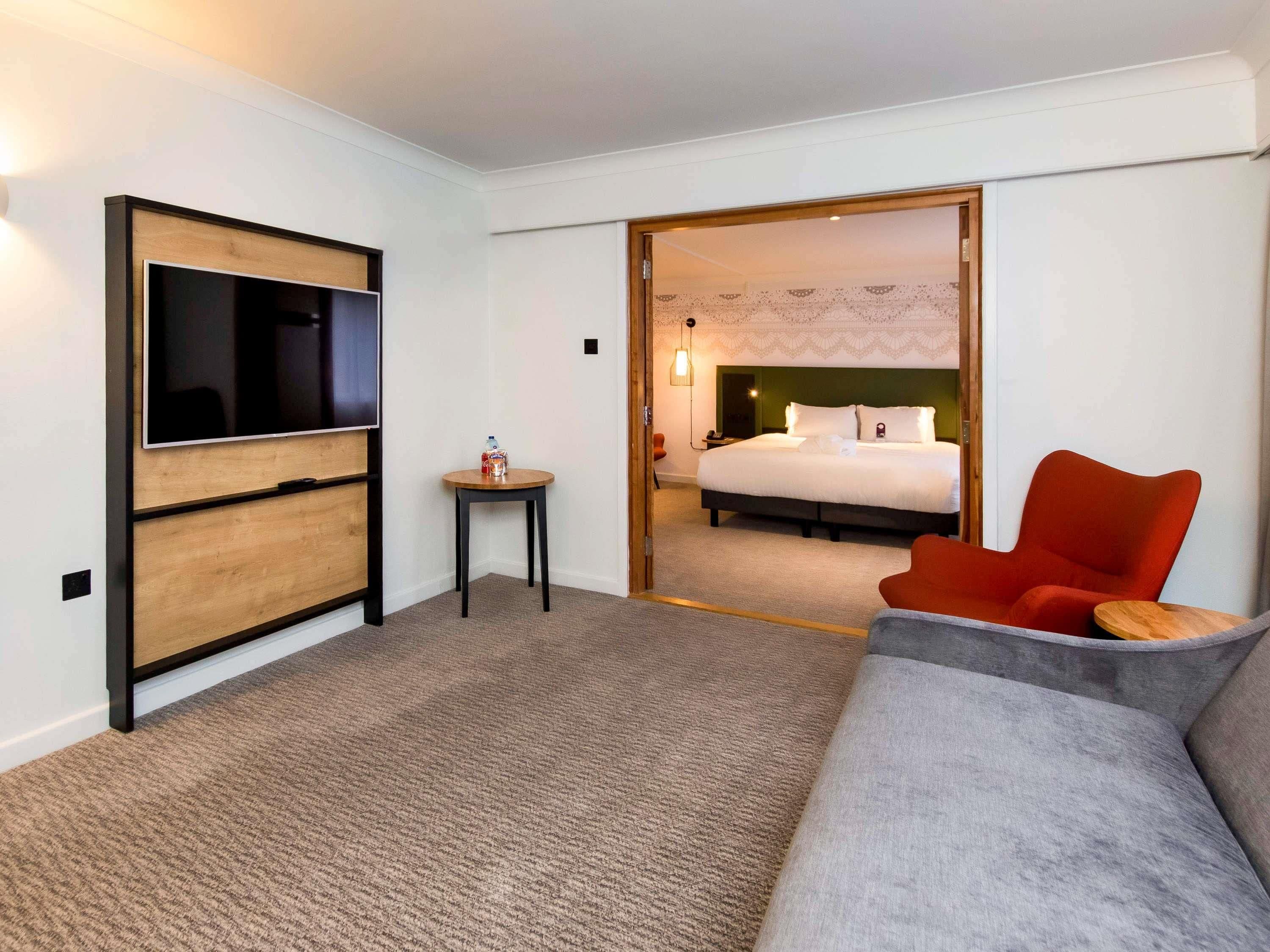 Habitación de hotel con televisor, una mesita con una botella, cama con almohadas y una silla roja.