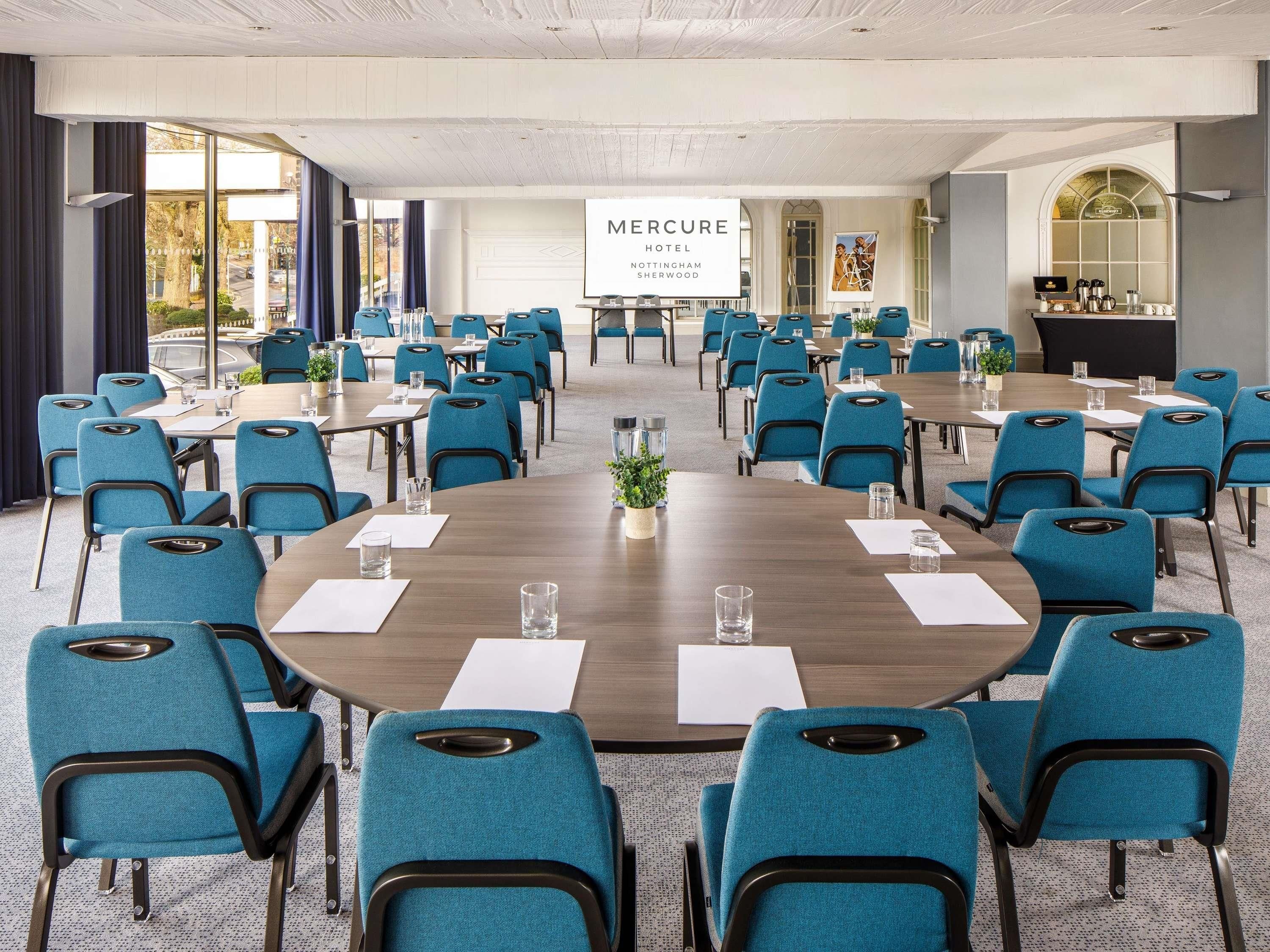 Ein Konferenzraum mit runden Tischen, blauen Stühlen und einem großen Banner mit der Aufschrift 'Mercure Hotel Nottingham Sherwood'.