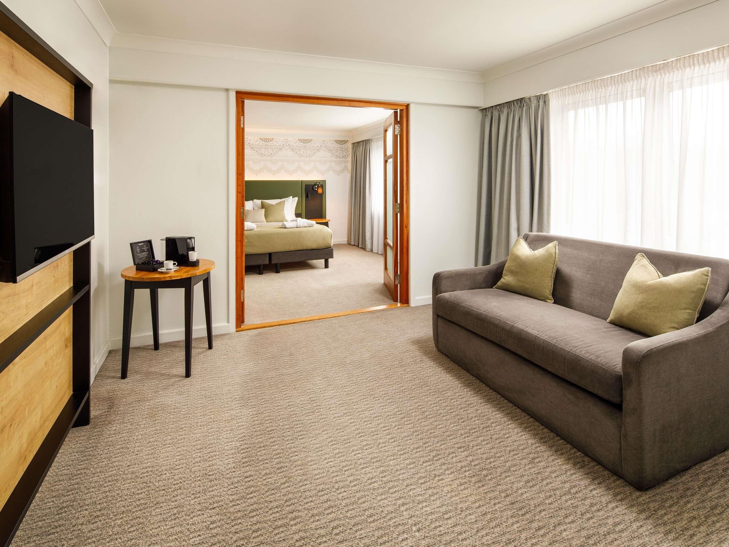 A hotel room with a large flat-screen TV, a sofa, a small table with a coffee maker, and a bed with a green headboard.