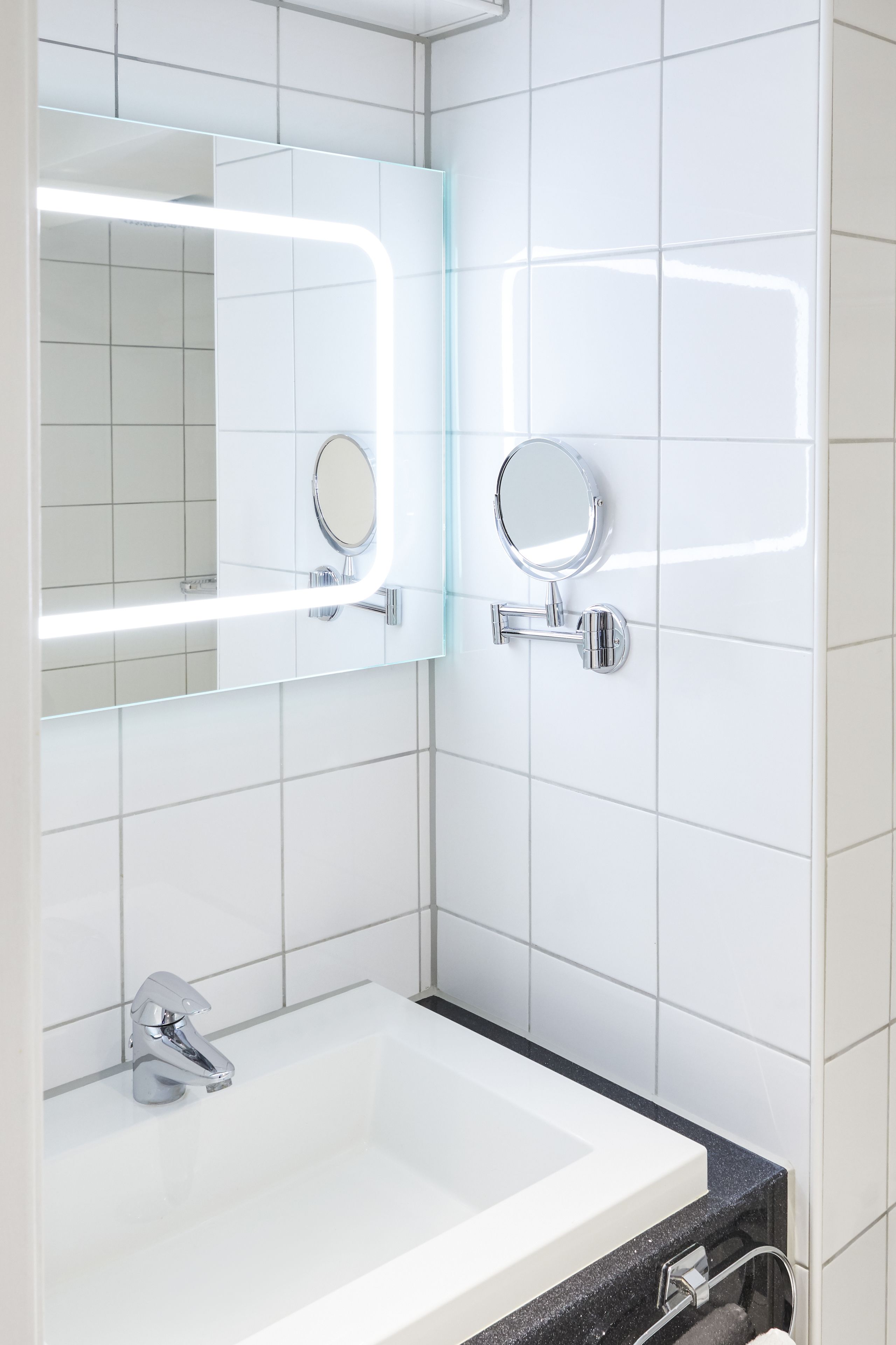 Un baño moderno con azulejos blancos, un lavabo blanco, un espejo con luz y un espejo de mano.