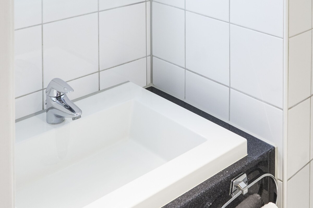 A modern bathroom with white tiles, a white sink, a mirror with a light, and a handheld mirror.