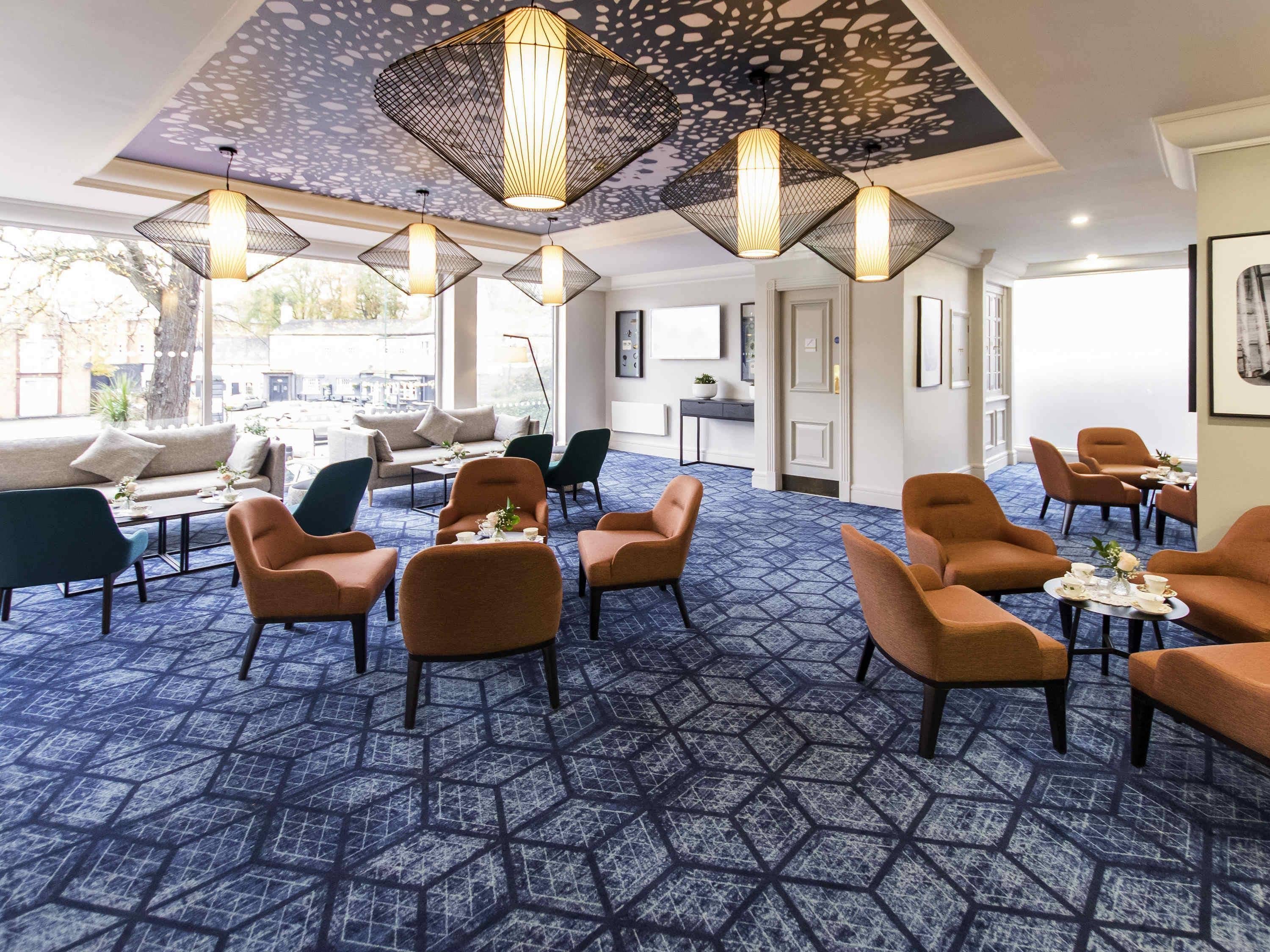 Egy hotel hallja, amelyben geometriai mintázatú szőnyeg, több ülősarok, kanapékkal és fotelokkal, valamint függesztett lámpatestek találhatók.