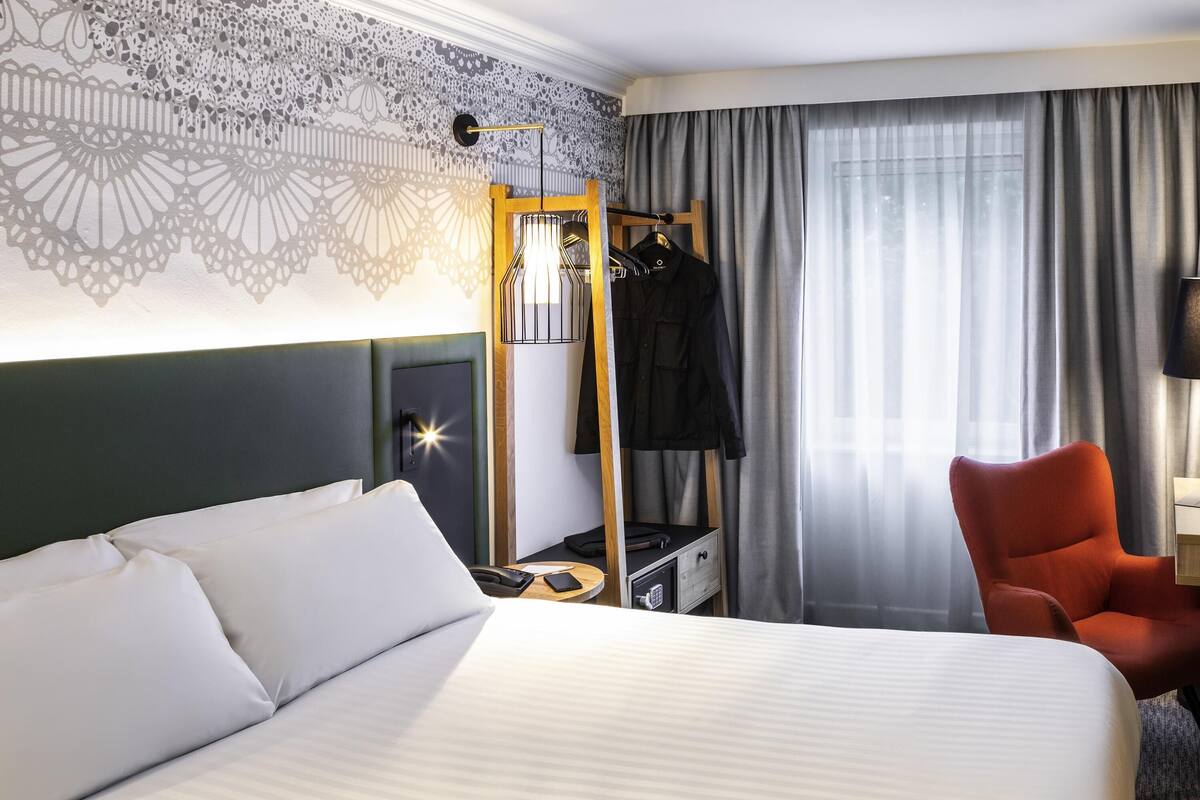 Une chambre d’hôtel avec un grand lit, un miroir, un porte-manteau, une chaise et une fenêtre avec des rideaux.