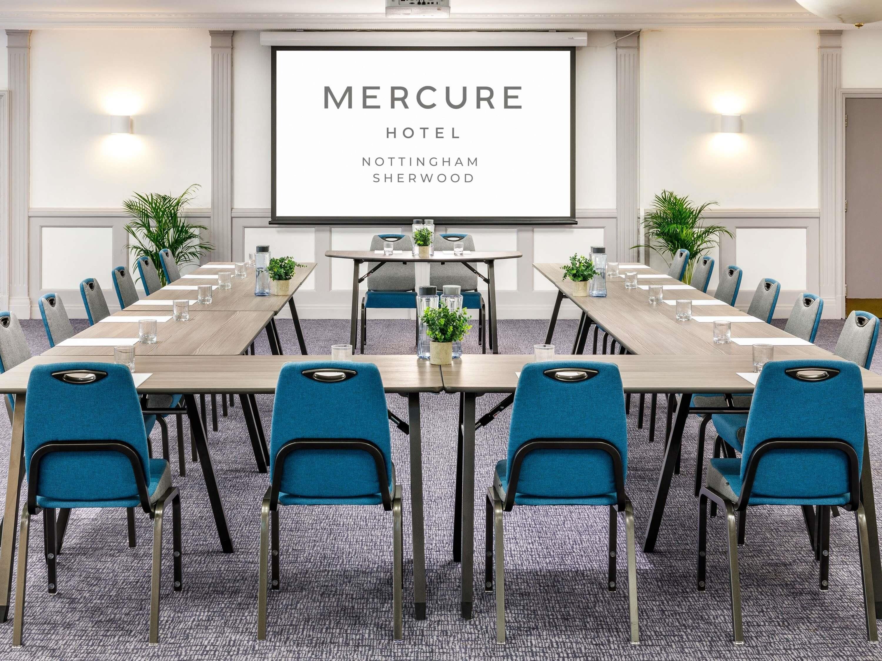 A conference room with a long table, blue chairs, and a screen displaying 'Mercure Hotel Nottingham Sherwood'.