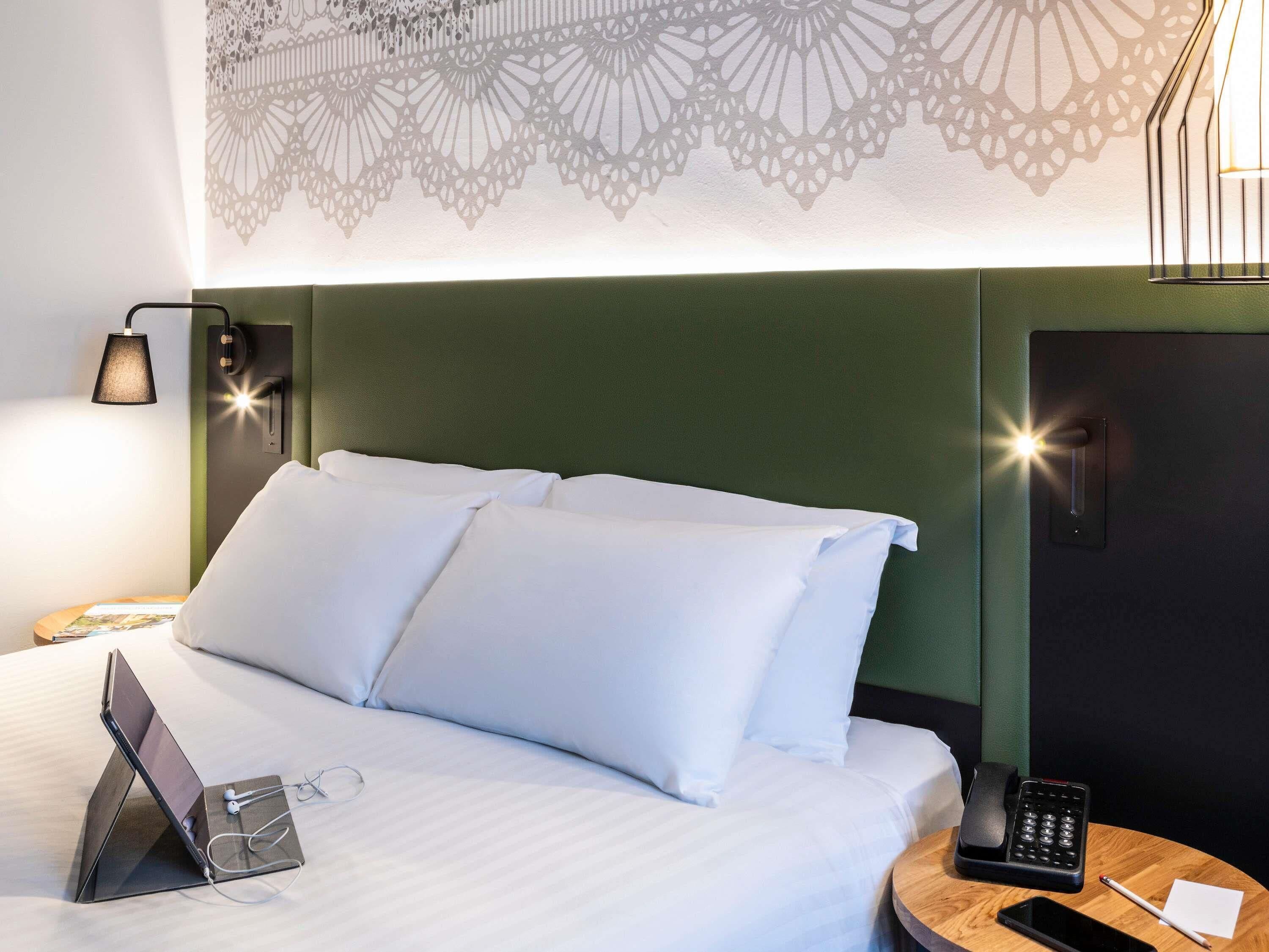 A neatly made bed with white linens, a laptop, and glasses on the bed, a bedside table with a phone and a notepad, and a wall with a geometric pattern.