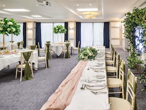 Un salón de banquetes con mesas dispuestas para un evento formal, con una mesa larga adornada con una tela rosa, sillas con cintas verdes y centros de mesa florales.