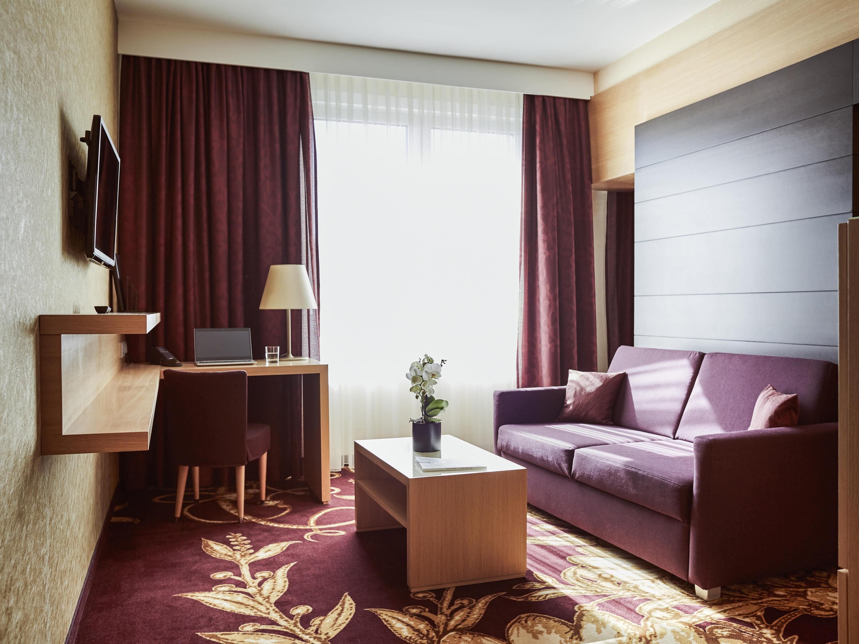 A hotel room with a desk, laptop, chair, sofa, and a plant.