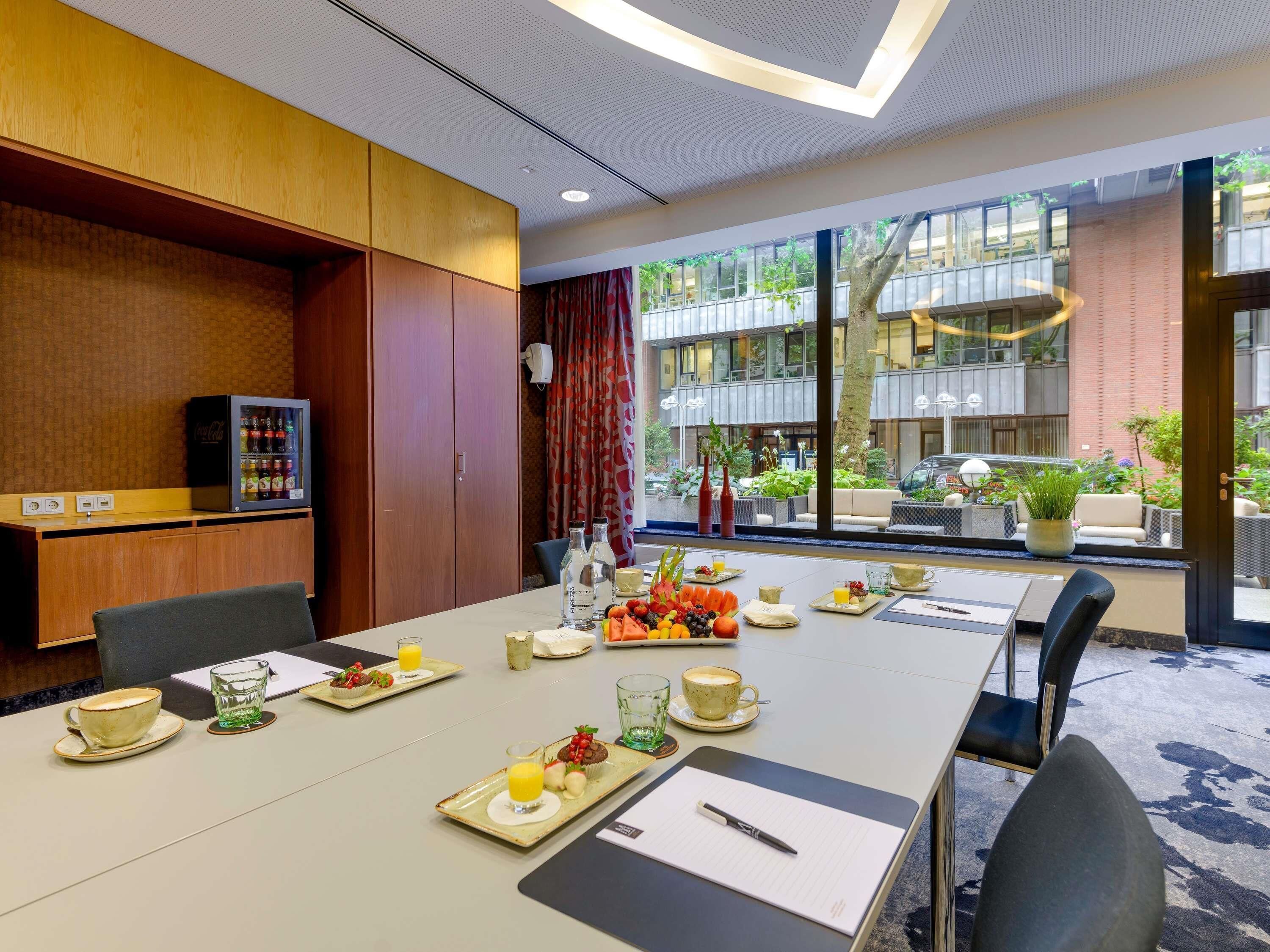 A conference room with a long table, chairs, a refrigerator with beverages, and a view of the outdoor area.