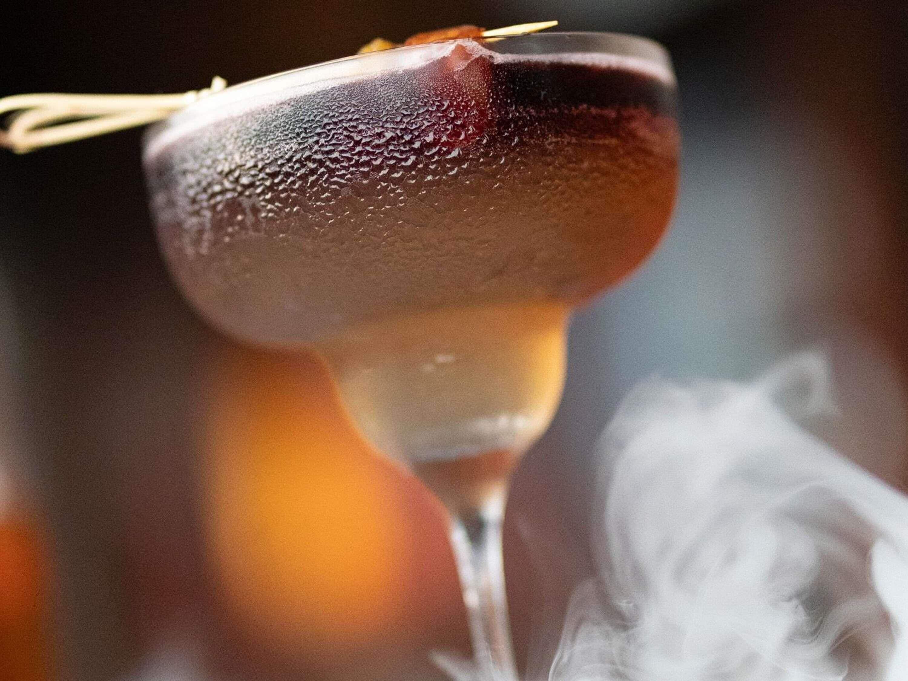 A close-up of a cocktail glass with condensation, a toothpick, and smoke.