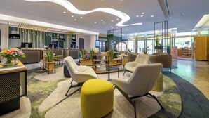 A modern hotel lobby with a reception desk, seating area, and a large window providing natural light. - Hotel Mondial am Dom Cologne - MGallery (Cologne)