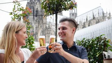 Deux personnes trinquent avec des verres de bière à une table en plein air, avec un bâtiment historique en arrière-plan.