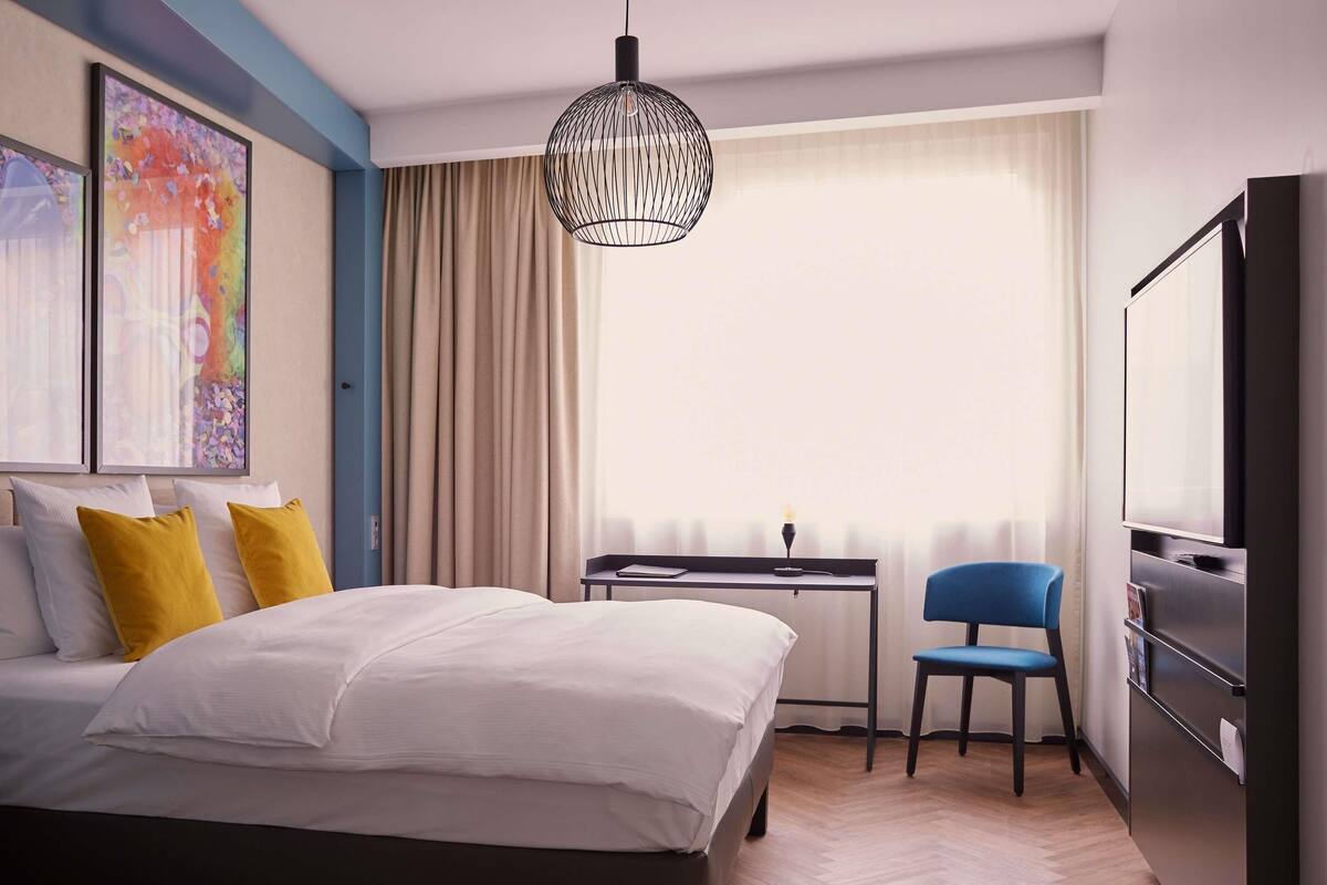 A hotel room with a bed, a desk, a chair, a television, and a large window with curtains.