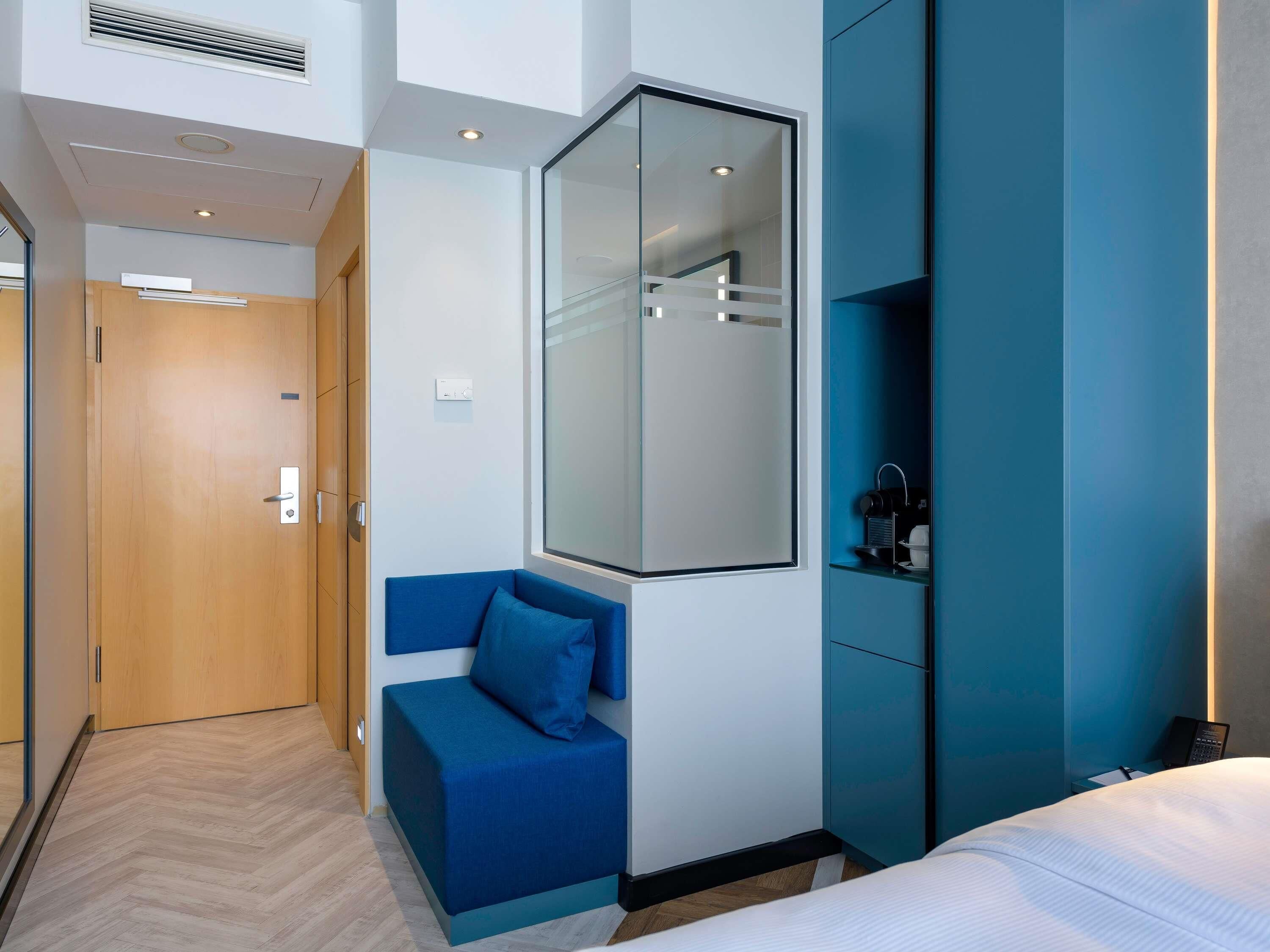 Une chambre d’hôtel moderne avec une porte en bois, un canapé bleu, un grand miroir et un lit avec une literie blanche.