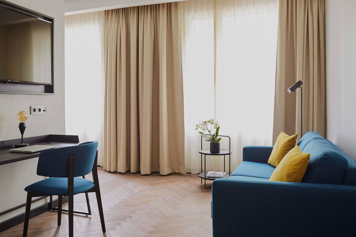 A modern living room with a blue sofa, a small side table with a plant, a desk with a lamp, and a large window with beige curtains.