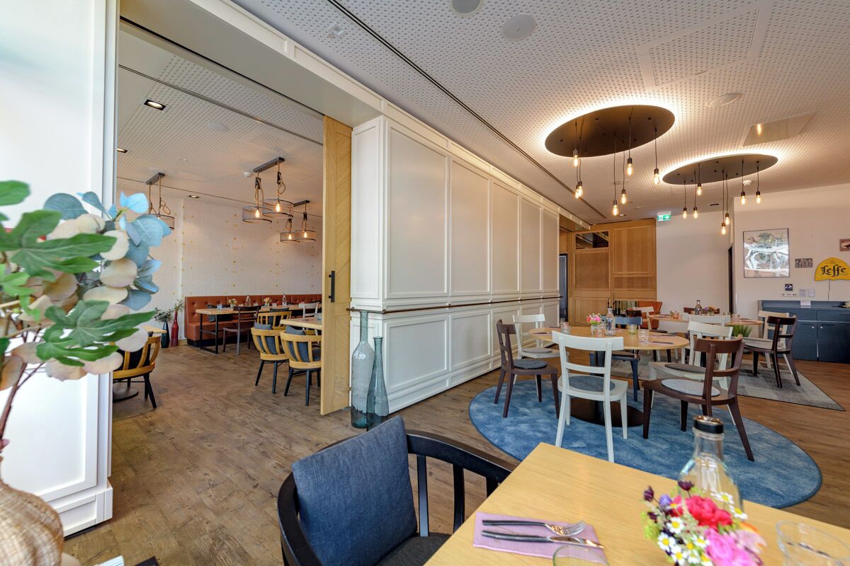 A modern restaurant interior with wooden flooring, white walls, and multiple dining areas.