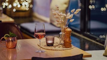 A cozy dining area with a wooden table, a glass of red wine, a copper mug, a small vase with dried flowers, and a candle on a woven placemat.