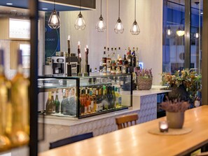 Zona de bar con una vitrina de vidrio llena de botellas, lámparas colgantes y una cesta de mimbre con flores.