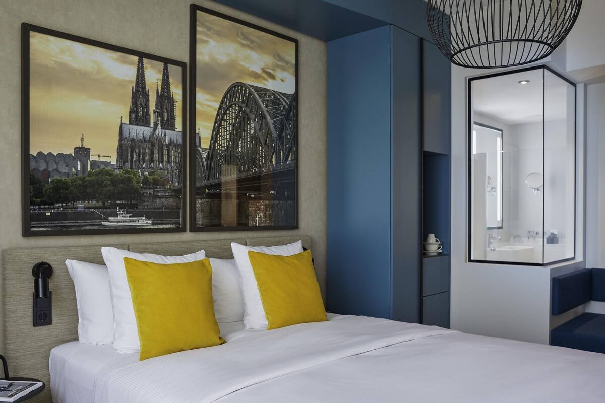 A hotel room with a bed, two framed pictures, a modern light fixture, and a bathroom with a mirror.