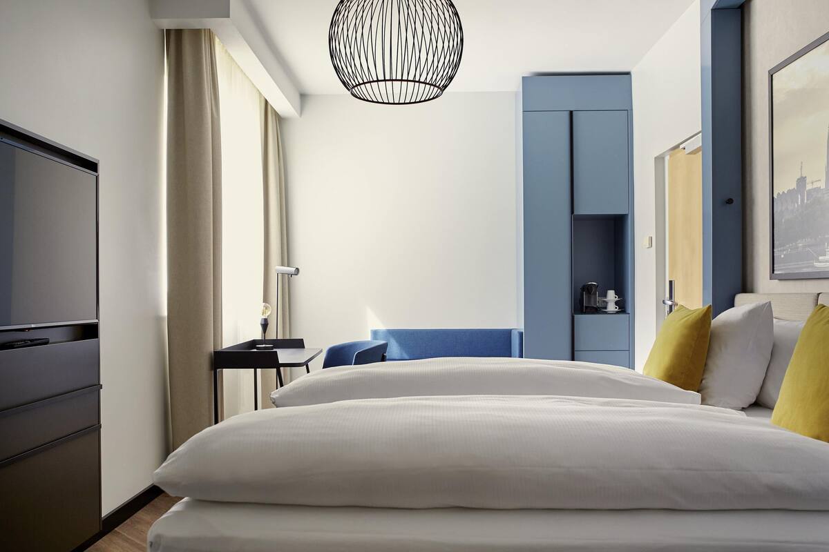 A hotel room with a bed, a desk, a chair, a television, a wardrobe, and a hanging light fixture.