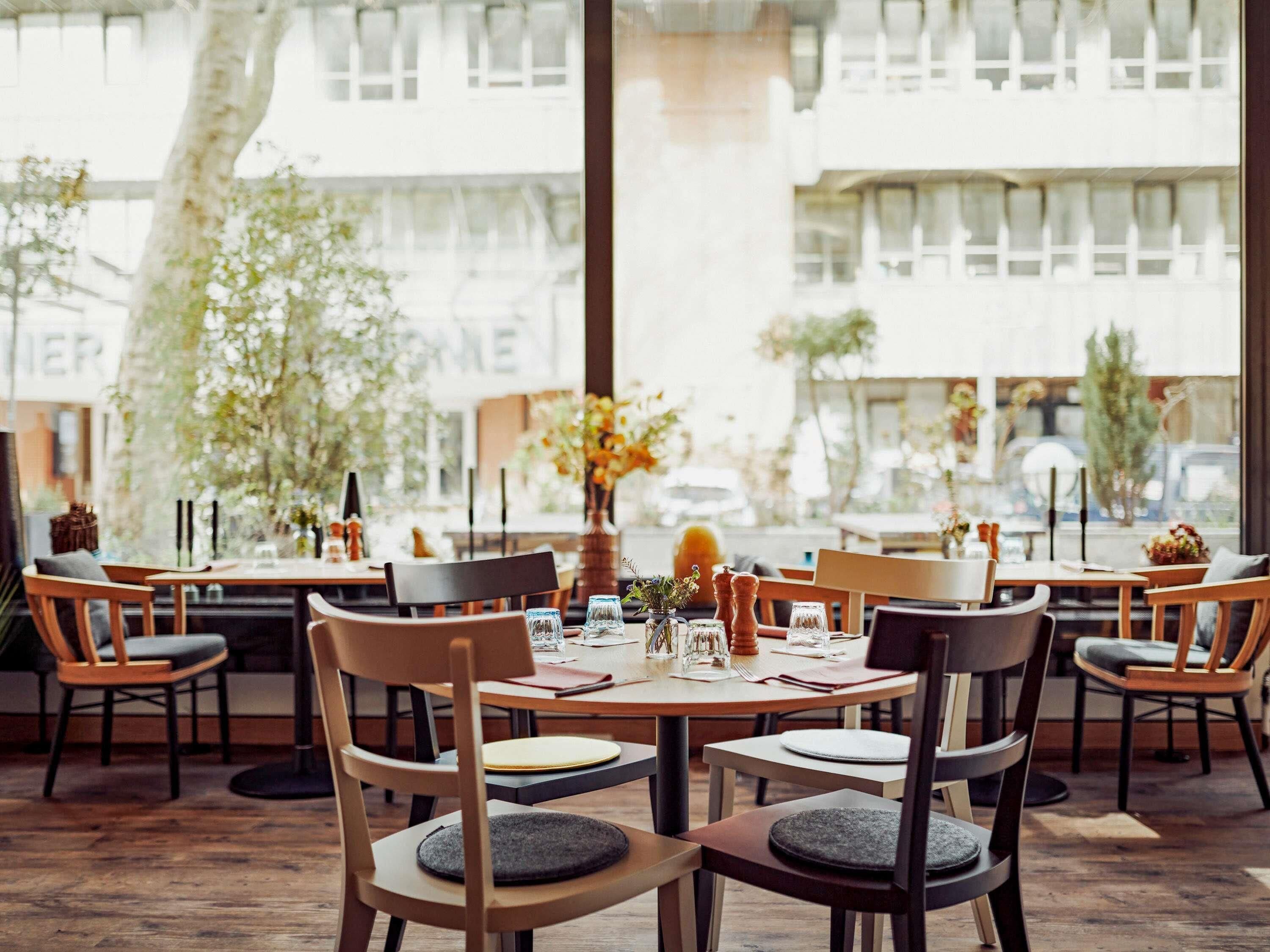 A restaurant with round tables and chairs, large windows, and indoor plants.