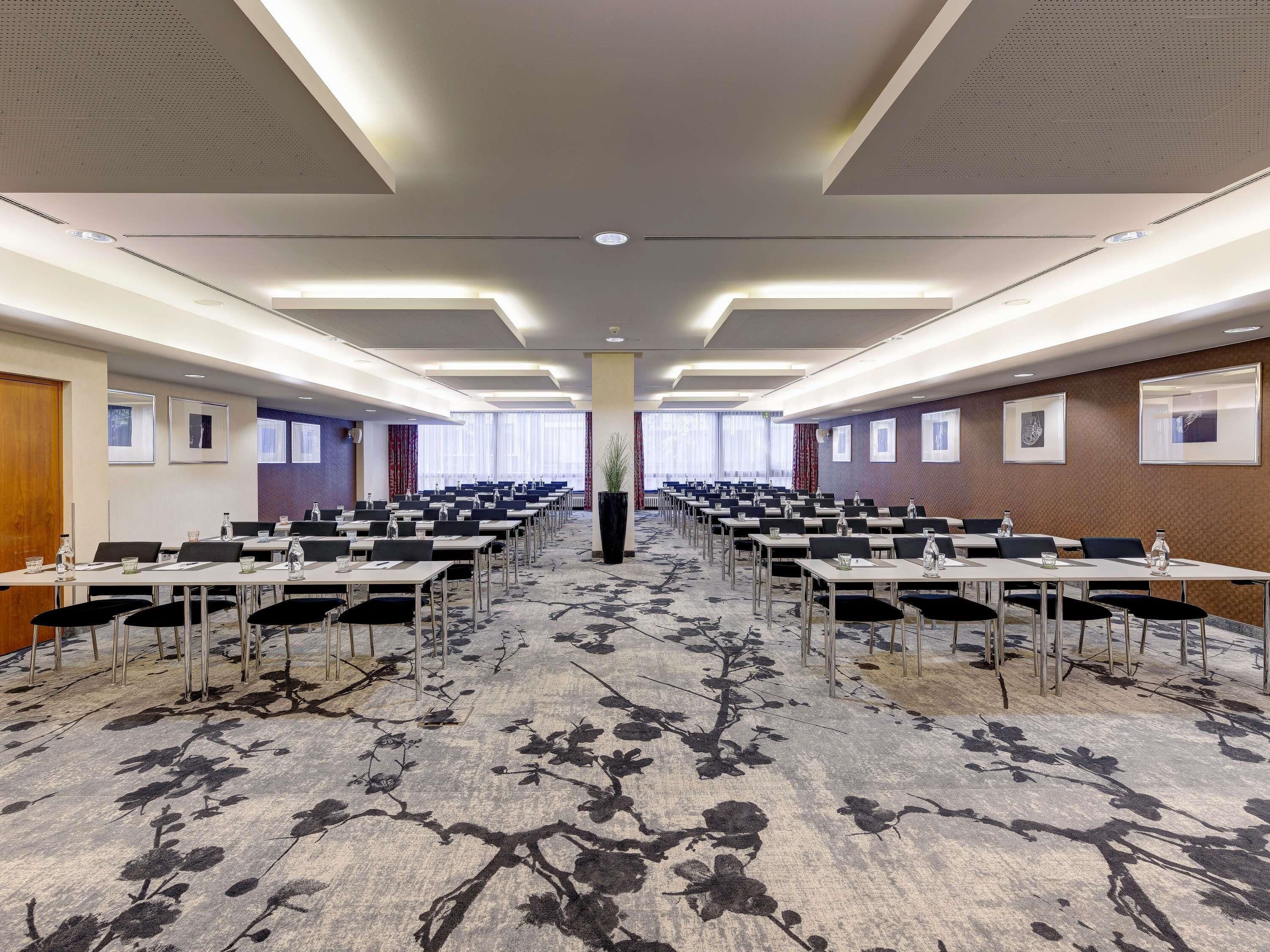 Une salle de conférence avec des rangées de tables et de chaises, une moquette à motifs et des tableaux encadrés accrochés aux murs.