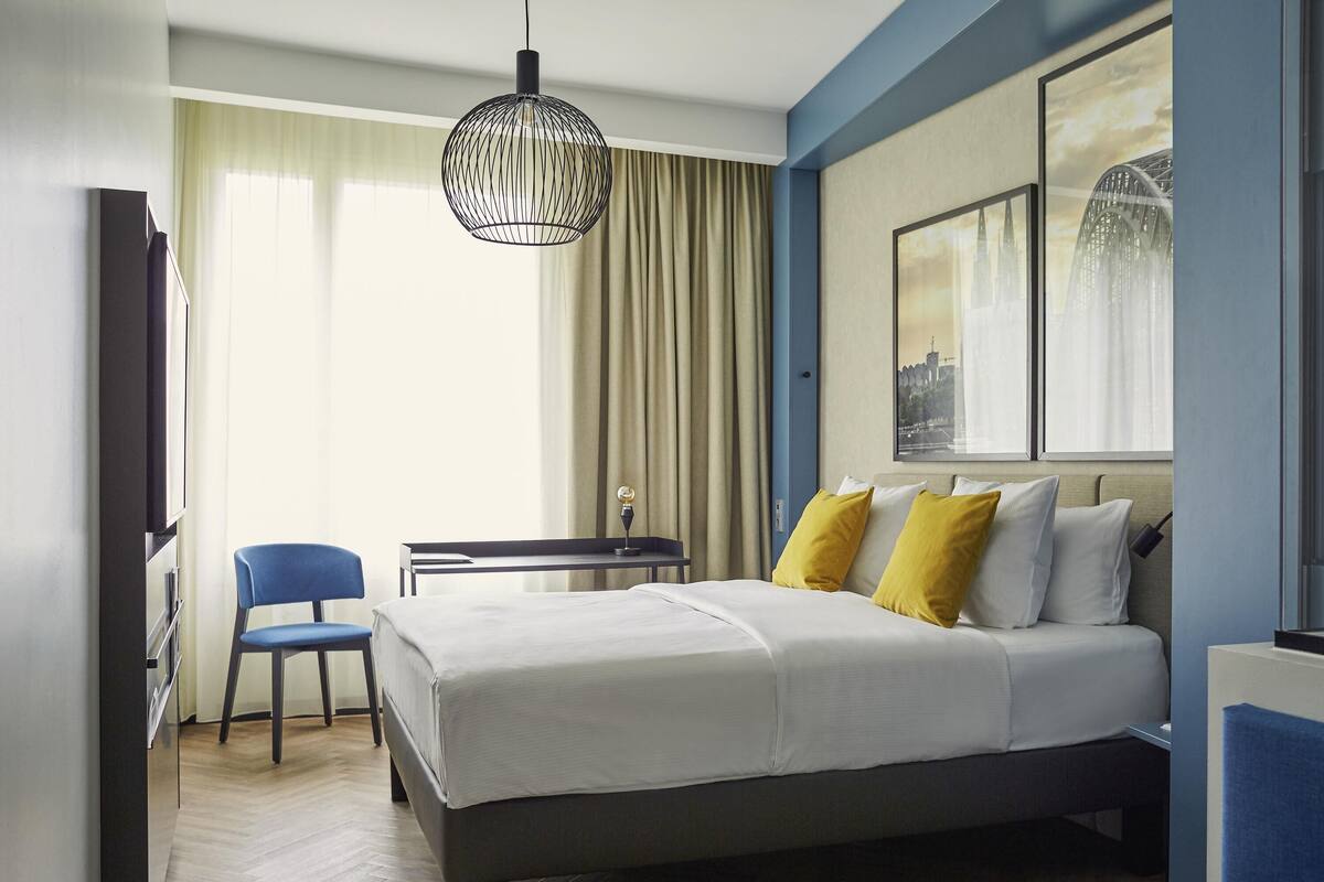 A hotel room with a bed, a desk, a chair, a television, and a large window with curtains.