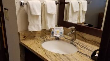 Un baño con lavamanos de mármol, dos toallas blancas colgadas en un perchero, un espejo y un cubo de basura.