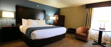 A hotel room with a large bed, two bedside tables with lamps, a small armchair, a round table, and a view of the outdoors.