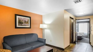 A hotel room with a gray sofa, a small wooden table, a framed picture on the wall, a floor lamp, and a carpeted floor.