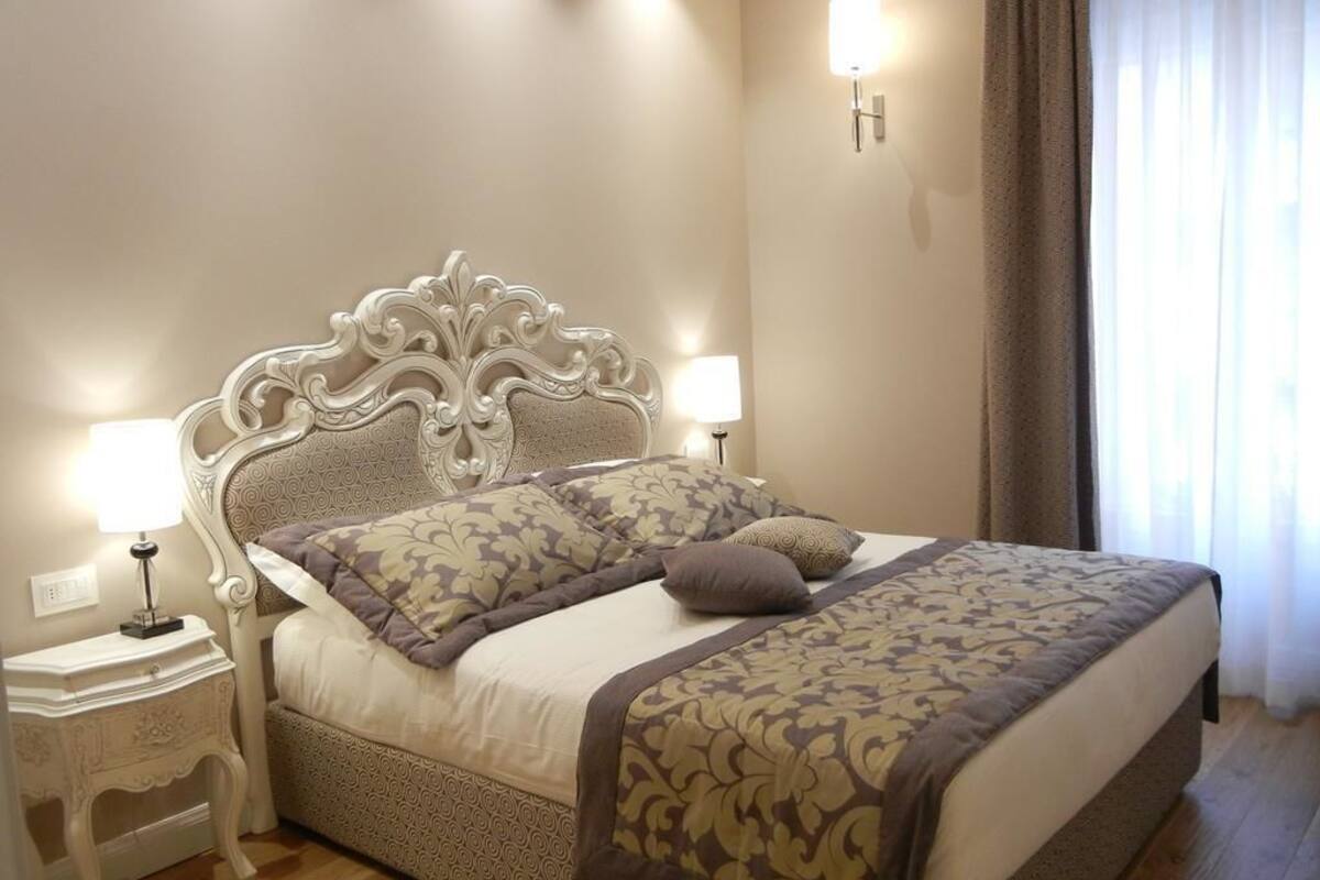 A neatly made bed with a patterned bedspread and pillows, a white ornate headboard, bedside lamps, and a nightstand.