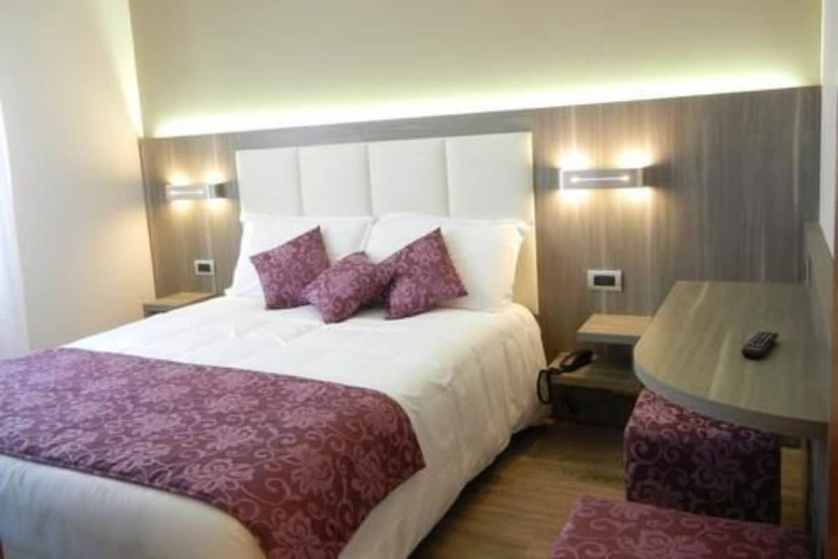 A hotel room with a large bed, two bedside tables, a round table, and a wall-mounted light fixture.