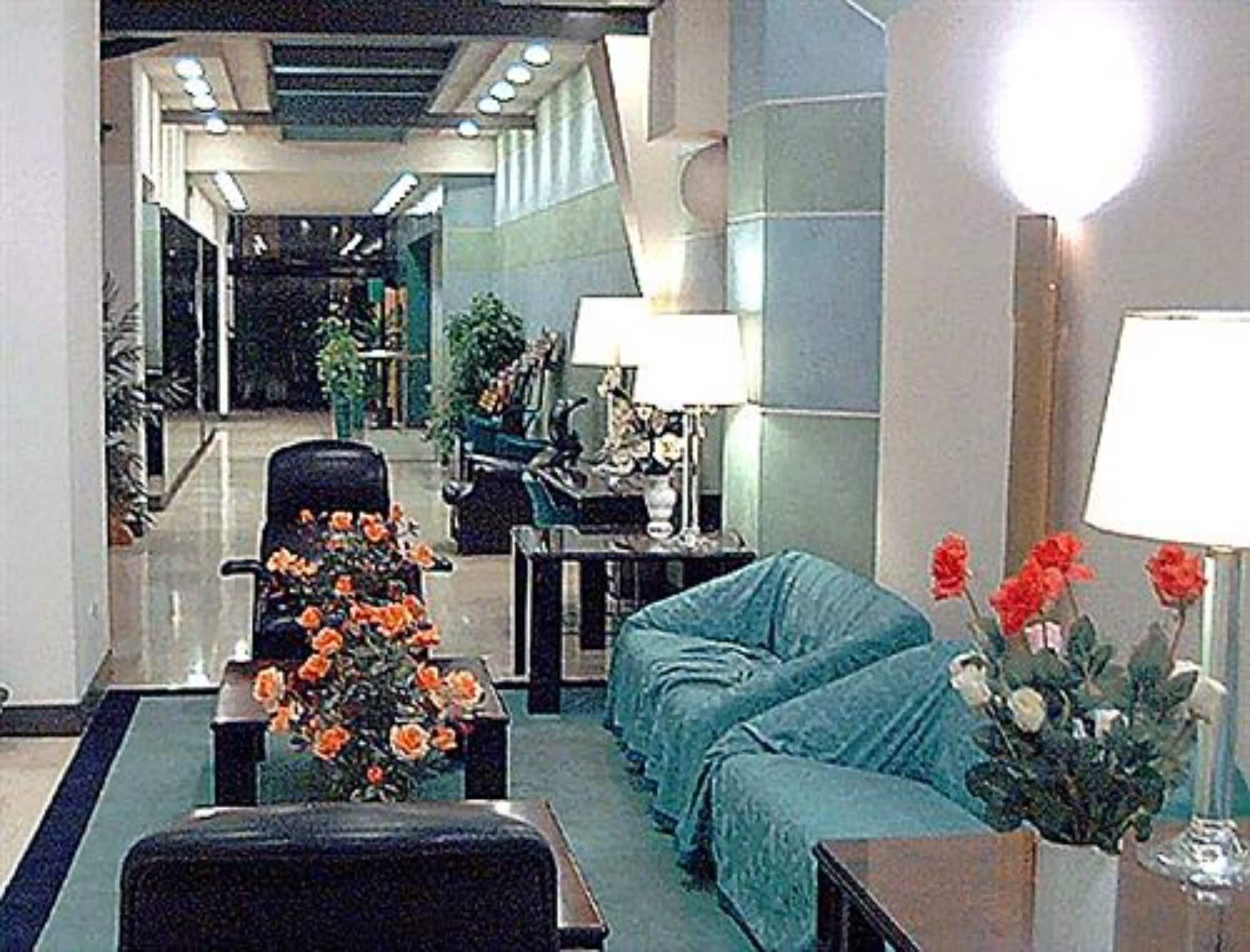 A lobby with a sofa, armchairs, a coffee table, and a vase of flowers. There is a lamp on a table and a chair in the background.