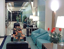 Ένας χώρος υποδοχής με έναν καναπέ, πολυθρόνες, ένα τραπεζάκι και ένα βάζο με λουλούδια. Υπάρχει ένα φωτιστικό πάνω σε ένα τραπέζι και μια καρέκλα στο βάθος.