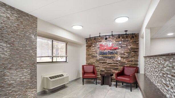 A room with a stone wall, two red chairs, a small table, and a window with blinds. - Red Roof Inn Greenville (Greenville)