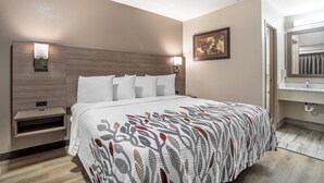 A hotel room with a bed, bedside tables, a wall-mounted lamp, a framed picture, and a bathroom with a mirror and sink. - Red Roof Inn Greenville (Greenville)