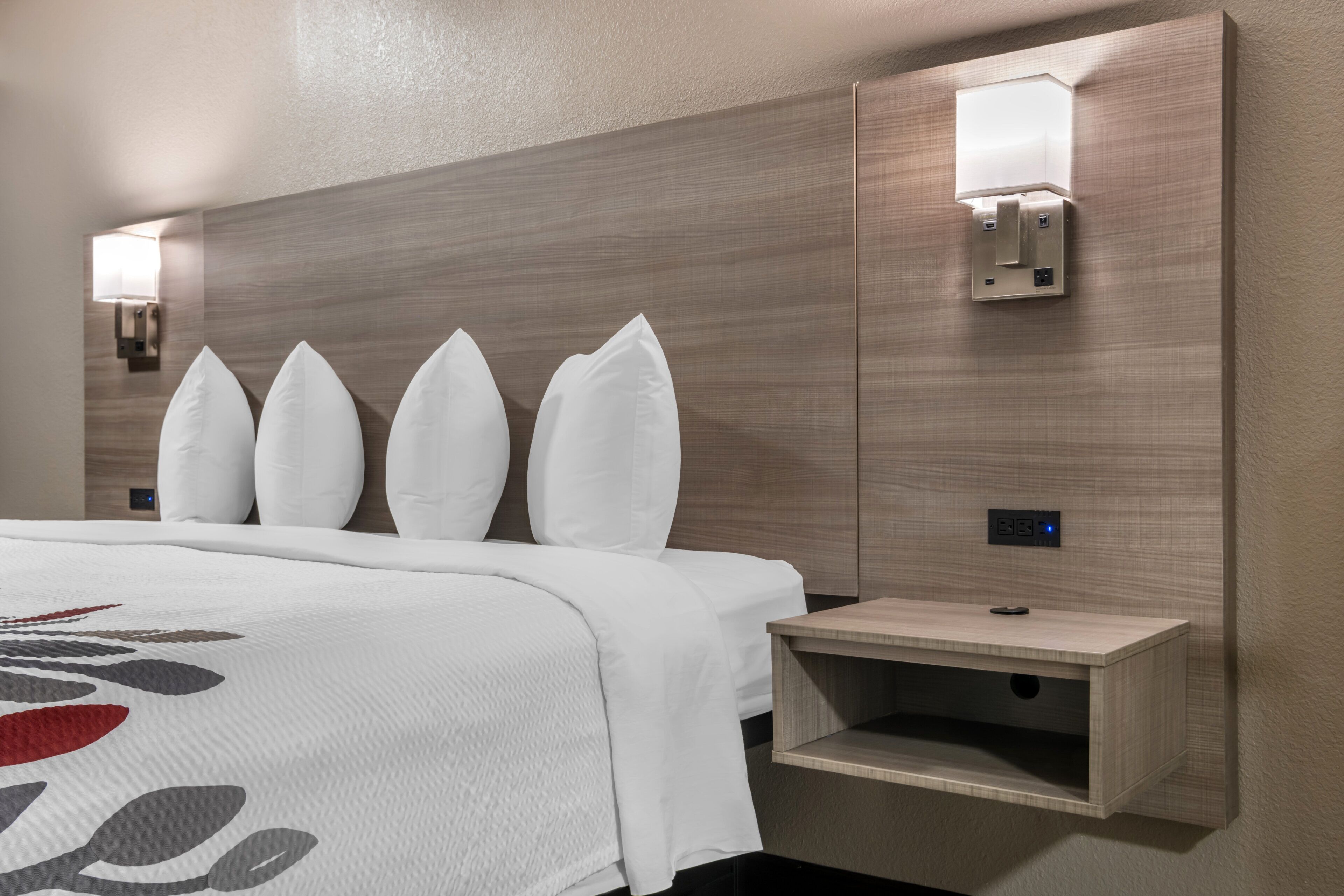 Una habitación de hotel con una cama, una mesita de noche, una lámpara de pared y un enchufe.