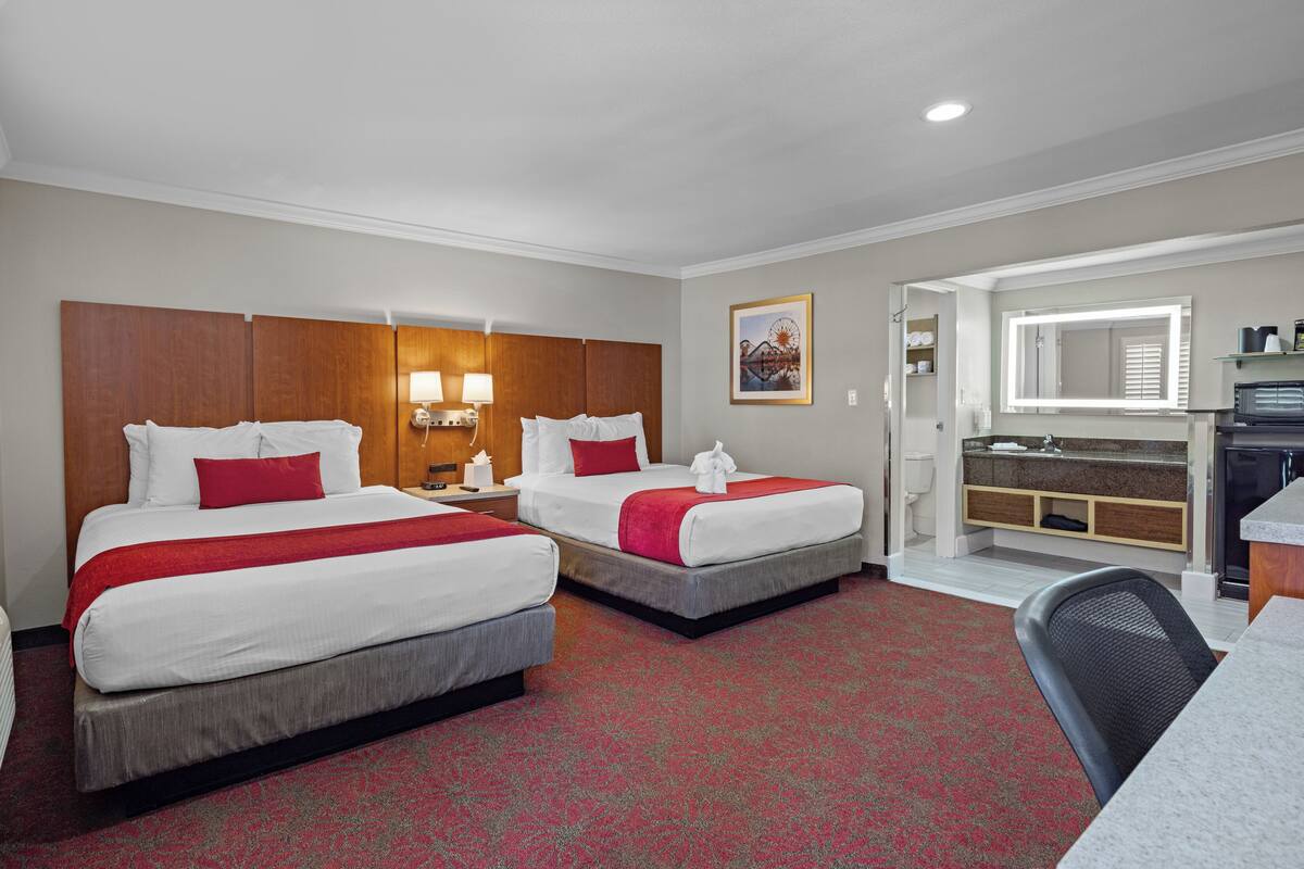 A hotel room with two beds, a desk, a chair, a bathroom with a mirror, and a small kitchenette.