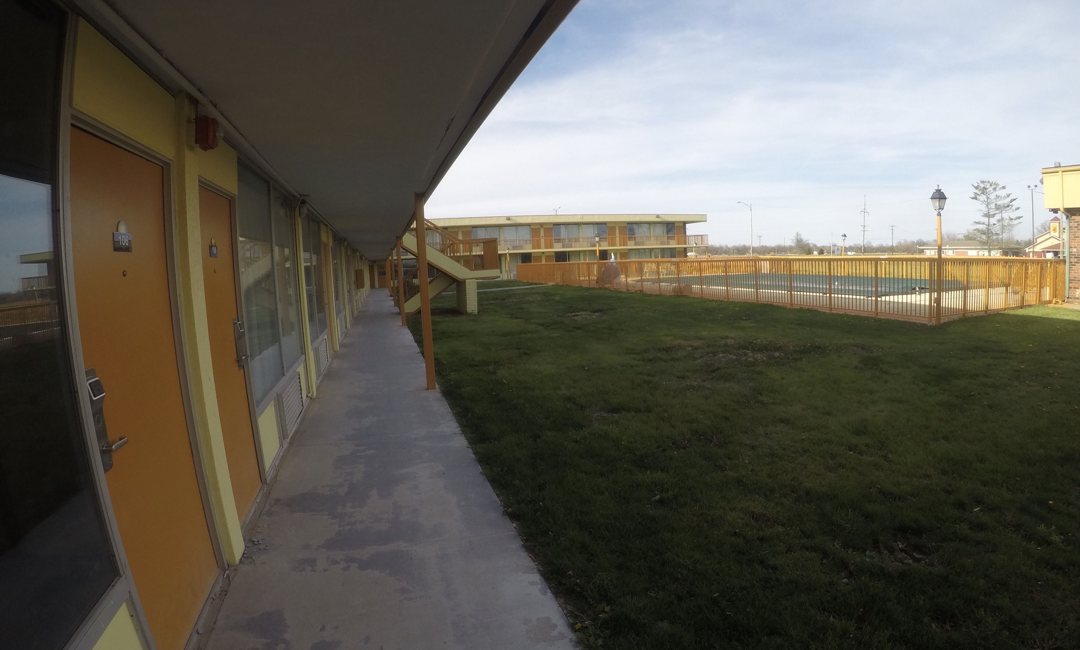 A motel with a covered walkway leading to a grassy area and a fenced pool.
