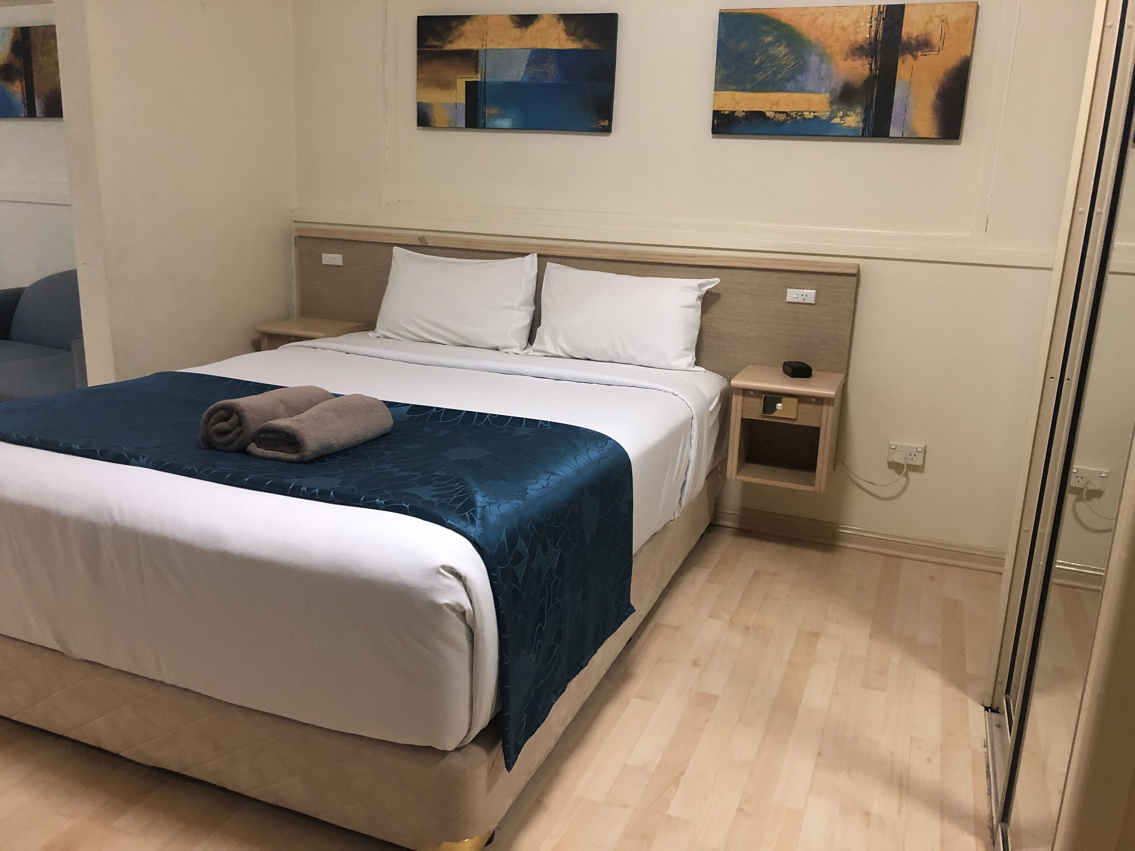 A neatly made bed with white and blue bedding, a wooden nightstand with a lamp, and artwork on the wall.