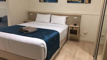 A neatly made bed with white and blue bedding, a wooden nightstand with a lamp, and artwork on the wall.