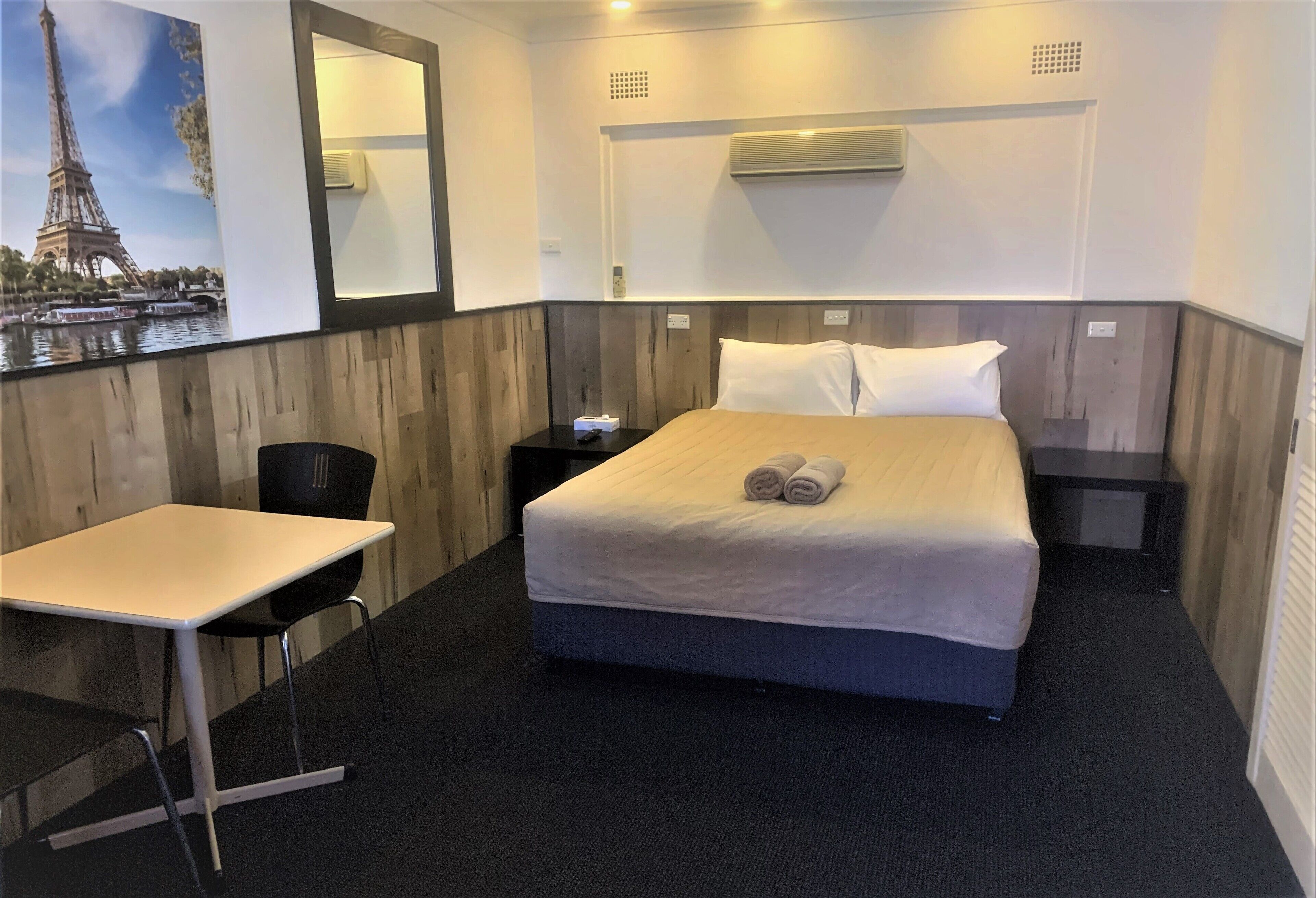 Habitación de hotel con cama, escritorio, silla, mesita de noche y aire acondicionado instalado en la pared.