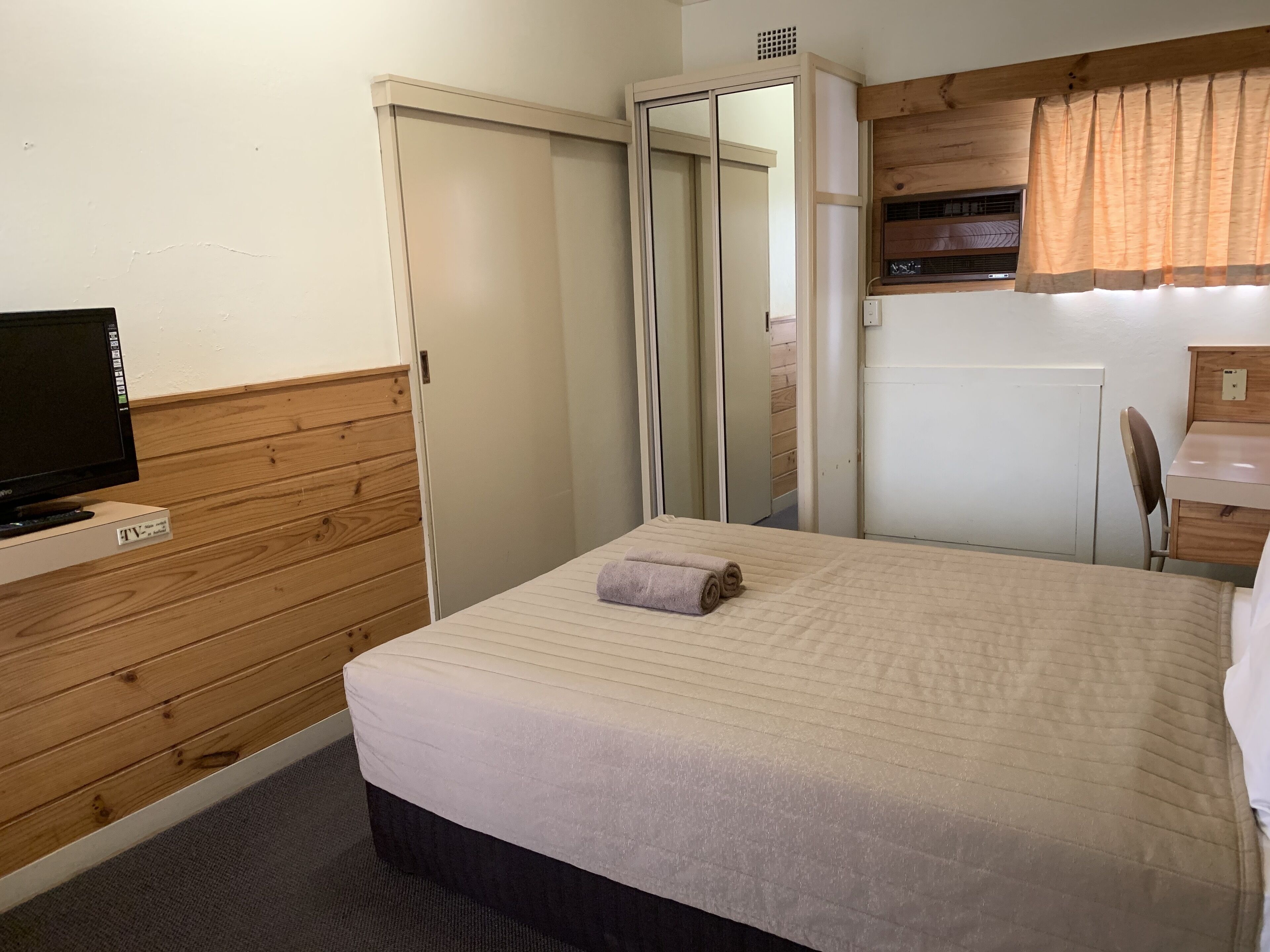 A hotel room with a bed, a TV, a wooden panel wall, a mirror, a desk, and a window with curtains.