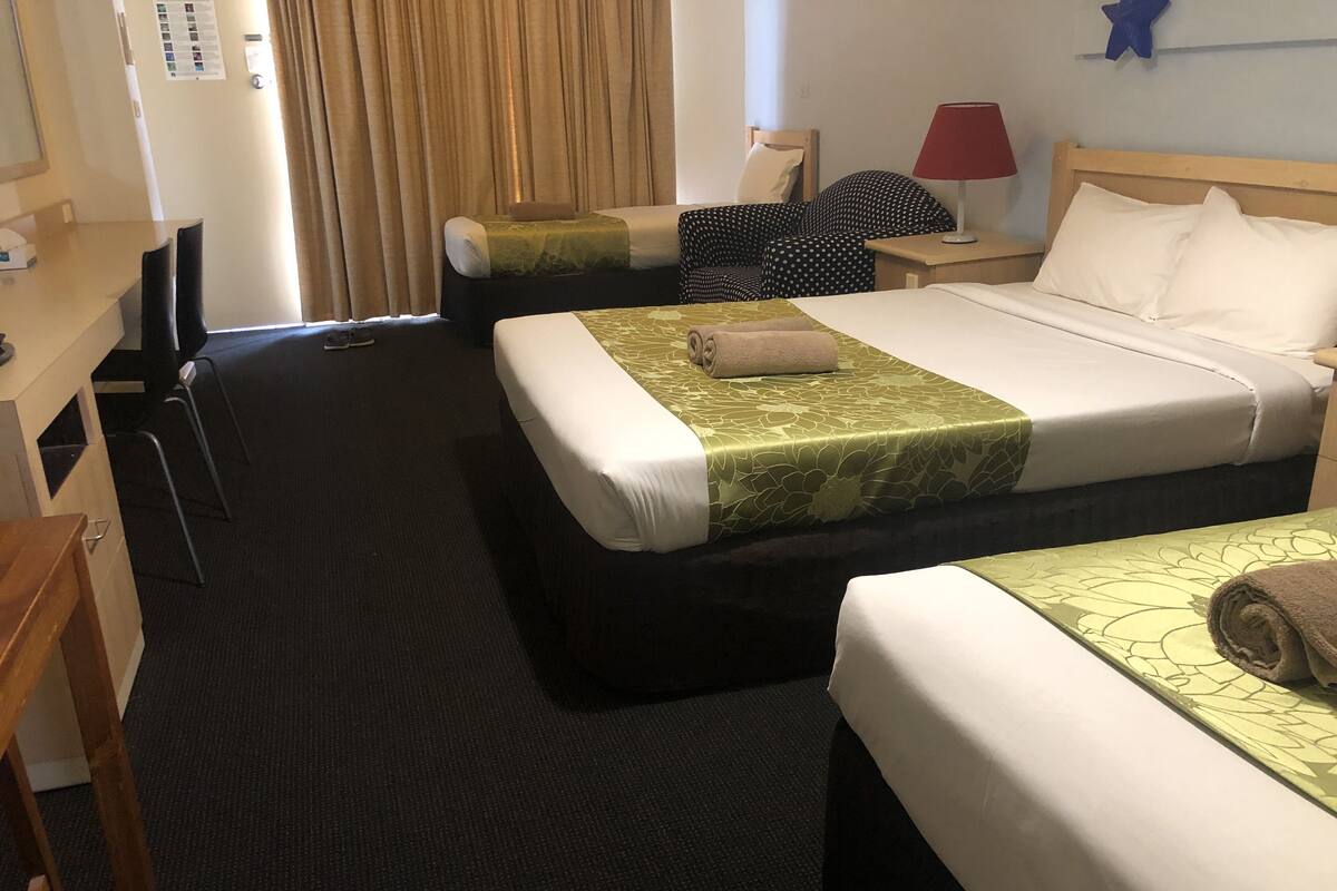 Ein Hotelzimmer mit zwei Betten, einem Schreibtisch, einem Stuhl, einem Nachttisch mit Lampe und einem Fenster mit Vorhängen.