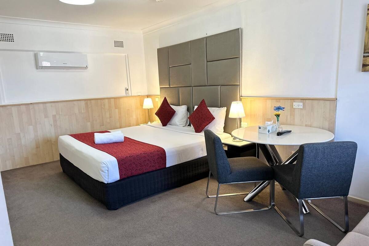 Ein Hotelzimmer mit einem Bett, zwei Nachttischen, einem runden Esstisch mit zwei Stühlen und einem modernen Kopfteil.
