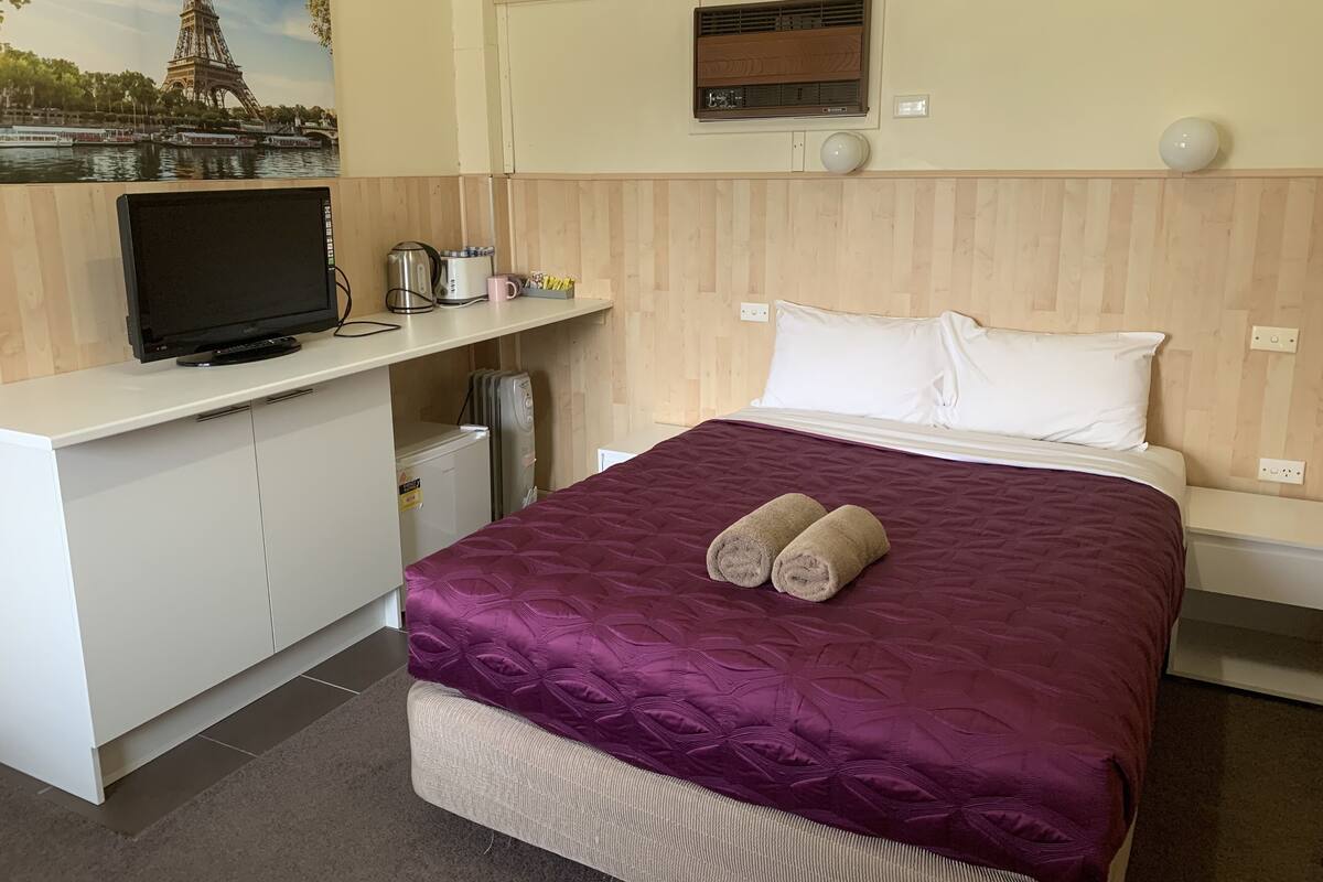 Ein Hotelzimmer mit einem Bett, einem Schreibtisch mit Fernseher, einer kleinen Kühlschrank und einem Gemälde des Eiffelturms.