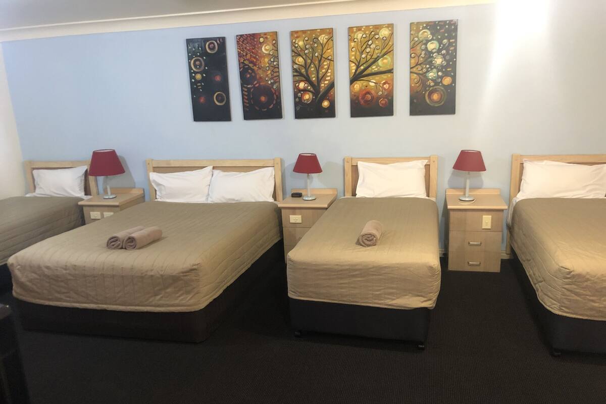 Ein Hotelzimmer mit vier Einzelbetten, jedes mit einem hölzernen Kopfteil und einer roten Lampe daneben. Darüber hängen drei Bilder an der Wand.