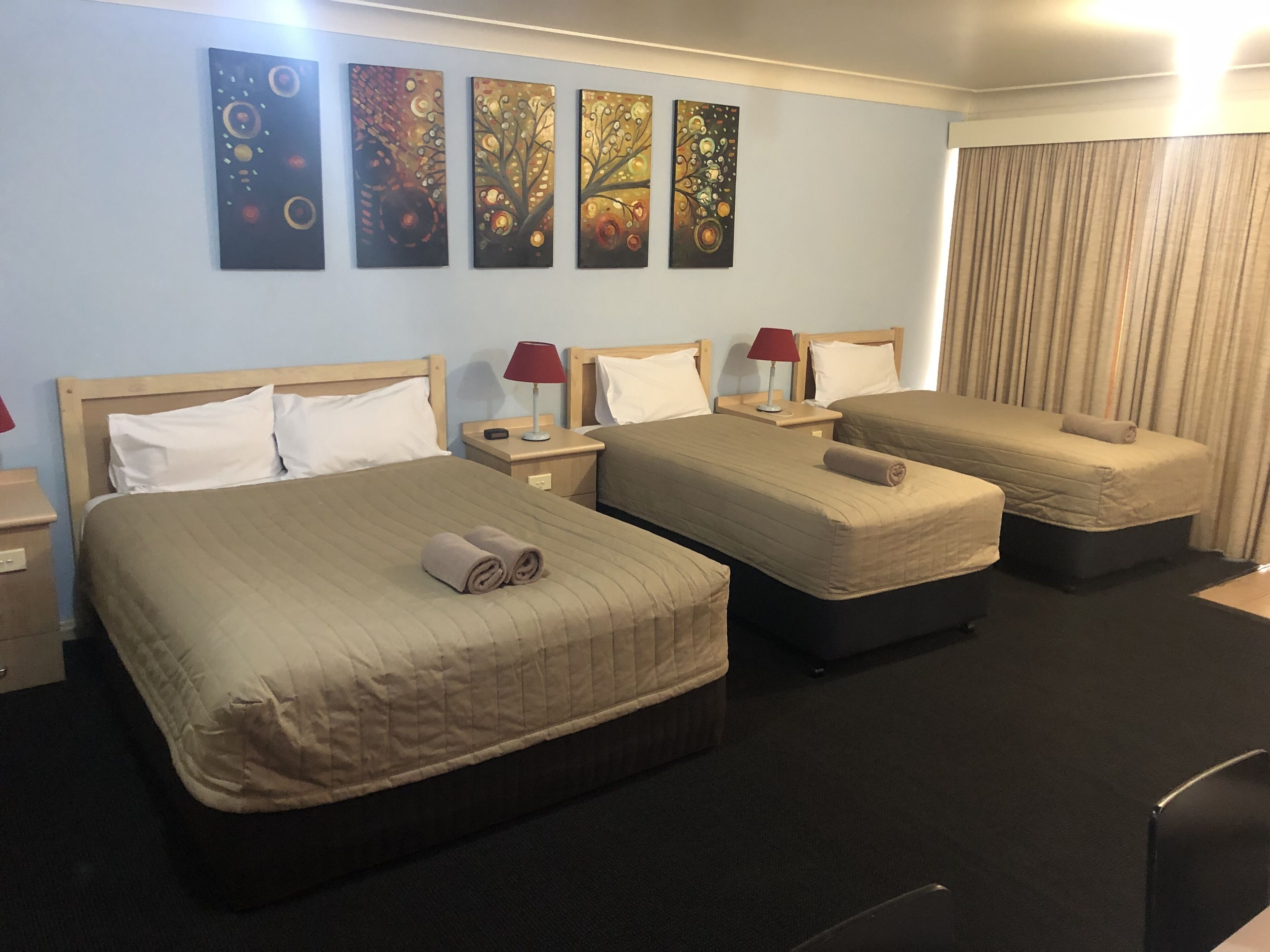 A hotel room with two single beds, a double bed, and a desk. There are four paintings on the wall, a red lamp on each bedside table, and a curtain on the window.